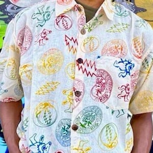 May include: Cream-colored button-down shirt with a colorful, repeating pattern of bears, lightning bolts, and other shapes. The shirt has short sleeves and a collared neckline. The shirt is worn by a person wearing sunglasses.