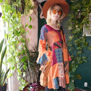May include: A long-sleeved tunic featuring a vibrant patchwork design with various orange, red, and blue patterned fabric panels. The garment has a V-neckline, flared sleeves, and a ruffled hemline, constructed from multiple contrasting textile sections.