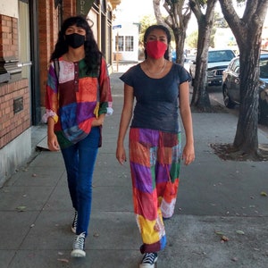 Puede incluir: Dos personas con mascarillas caminan por una acera. Una persona lleva una camisa de retazos con vaqueros azules y zapatillas negras. La otra persona lleva una camiseta azul oscuro, pantalones de retazos y zapatillas negras. Hay árboles y coches aparcados en el fondo.