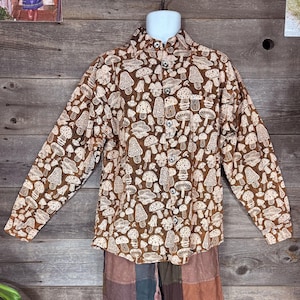 Puede incluir: Camisa marrón con botones con un estampado repetido de setas blancas. La camisa de manga larga se muestra en un maniquí. Tiene cuello y botones en la parte delantera. La tela parece ser un material suave y ligero.