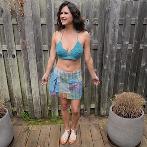 Op de afbeelding: Een turquoise gehaakte haltertop en een patchwork minirok. De rok heeft vierkanten stof in blauw, groen, paars en geel. De outfit wordt gecombineerd met witte sandalen met bruine zolen. De achtergrond is een houten hek en potplanten.