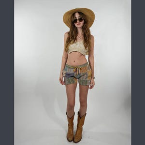 May include: A woman wearing a straw hat, sunglasses, a crocheted top, patchwork shorts, and brown cowboy boots. The shorts have a drawstring waist and a mix of colors including green, yellow, and purple.