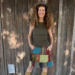 May include: A woman wearing an olive green ribbed tank top and a patchwork mini skirt with various colors and patterns. She is standing in front of a weathered wooden wall, with her hands in her pockets.