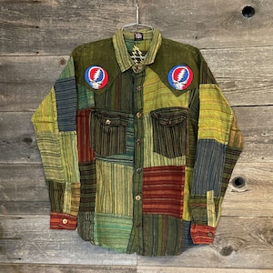 May include: A patchwork long-sleeve shirt with a green, brown, and red color scheme. The shirt has two embroidered patches on the chest, each featuring a circular design with a skull and lightning bolt.