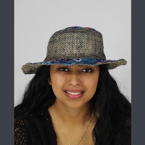 Könnte beinhalten: Ein gewebter Strohhut mit einem gemusterten Stoffband. Der Hut hat eine breite Krempe und ein neutrales Farbschema mit einem blau, orange und türkis gemusterten Band um den Kopf. Der Hut wird von einer Person mit langen schwarzen Haaren getragen.