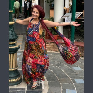 May include: A woman wearing a patchwork print maxi dress with a matching shoulder bag. The dress features a vibrant mix of red, blue, and green patterns. The bag has a long strap and a similar patchwork design. The woman is wearing silver sandals.