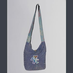 May include: A tie-dye shoulder bag in shades of blue with a colorful dancing bear applique. The bag has a long strap with floral patterns in pink and blue. The bag is made of soft fabric.