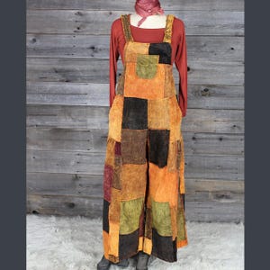May include: A patchwork overall dress in shades of orange, brown, black, and green. The dress has a square neckline, wide straps, and a large front pocket. A long-sleeved red shirt is worn underneath, and a scarf is draped around the neck.