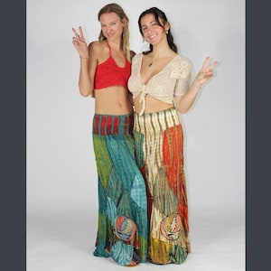 Op de afbeelding: Twee vrouwen in kleurrijke bohemien outfits. De ene draagt een rode haltertop en een patchwork rok met een Grateful Dead logo. De andere draagt een crèmekleurige gehaakte top en een vergelijkbare patchwork rok. Beiden lachen en geven vredestekens.