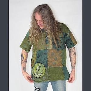 May include: Green and brown patchwork shirt with a round patch featuring a skull and lightning bolt design. The shirt has short sleeves and a button-up neckline. The shirt is worn with blue jeans.