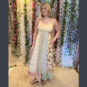 May include: A long, flowing dress with a patchwork design. The bodice is a light yellow with thin straps. The skirt features panels of various patterns, including floral and paisley prints, in shades of cream, green, and pink. The dress is worn with sandals.