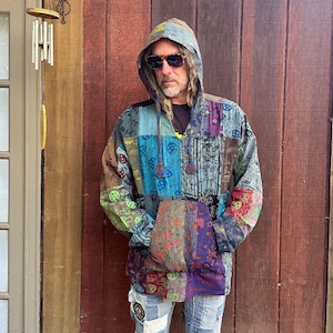 Peut inclure: Un pull à capuche à manches longues présentant un motif patchwork avec divers imprimés colorés, incluant des dessins de champignons. Le vêtement comprend une poche kangourou à l'avant, une capuche à cordon et des tons bleus, violets et verts.