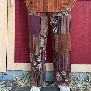 Può includere: Pantaloni patchwork con una varietà di motivi e colori, tra cui marrone, viola e tessuti fantasia. I pantaloni hanno un girovita con coulisse e sono indossati con una camicia a righe abbottonata. I pantaloni sono fatti di più toppe di tessuto.