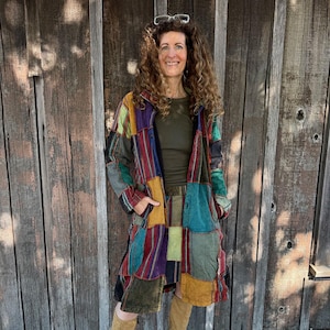 Puede incluir: Un abrigo patchwork multicolor, con paneles rojos, azules, verdes y amarillos. El abrigo largo se lleva sobre un top y una falda verdes. El fondo es una pared de madera envejecida.