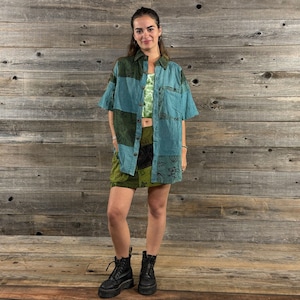May include: A woman wearing a blue and green patchwork shirt with a short sleeve and a green and black patchwork skirt. She is wearing black boots.