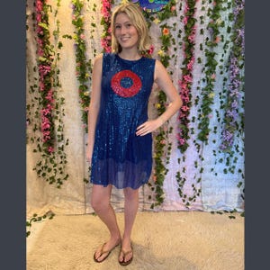 May include: A sleeveless, knee-length blue sequin dress with a red circle design. The dress has a round neckline and a loose, flowing fit. The model is wearing gold-coloured sandals.