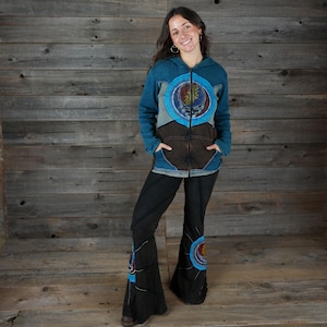 Peut inclure: Un sweat à capuche zippé bleu sarcelle et marron avec un motif circulaire, associé à un pantalon évasé noir. Le sweat à capuche a une capuche et des poches. Le pantalon a un motif assorti sur le bas évasé. L'ensemble est porté sur un fond en bois.