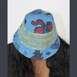 Könnte beinhalten: Ein blauer Fischerhut mit einem gemusterten Design mit einer roten und schwarzen Kreatur, schwarzen Bäumen und anderen Symbolen. Der Hut hat ein gewebtes Band um den Rand.