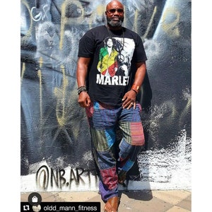 May include: A man wearing a black t-shirt with a graphic of Bob Marley and the text "MARLEY" on the front. He is also wearing colorful patchwork pants and sandals.