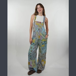 May include: A patchwork print jumpsuit with wide legs. The jumpsuit features a mix of floral and geometric patterns in shades of blue, yellow, and green. It has a bib-style top with adjustable straps and pockets.