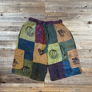 May include: Patchwork shorts with a variety of colors including green, blue, brown, and purple. The shorts feature multiple square patches with Grateful Dead bear and lightning bolt designs. They have two cargo pockets and an elastic waistband.