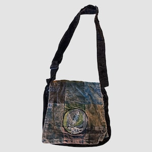 Könnte beinhalten: Schwarze Cord-Umhängetasche mit Patchwork-Design in Blau-, Grün- und Brauntönen. Die Tasche hat einen runden Aufnäher mit einem Totenkopf- und Blitzmotiv. Der verstellbare Riemen ist ebenfalls schwarz.
