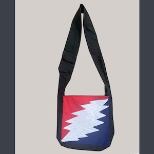 May include: Black shoulder bag with a red, white, and blue lightning bolt design on the front. The bag has a long black strap and is made of a durable fabric. The design is a stylized lightning bolt.