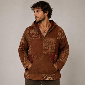 Puede incluir: Una sudadera con capucha marrón que presenta un diseño de patchwork con varios patrones intrincados, incluido un motivo de árbol de la vida en el pecho. La prenda tiene mangas largas, un bolsillo canguro delantero y un forro interior rojo oscuro.