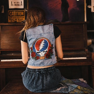 May include: A light blue denim vest featuring a large embroidered patch on the back. The patch displays a white skull with a blue and red lightning bolt design, surrounded by red roses. The vest is worn over a black short-sleeved shirt.