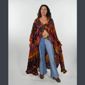 May include: A long, flowing tie-dye kimono in shades of orange, pink, and black, worn open over two-tone denim bell-bottom pants. The outfit is accessorized with a brown belt featuring a gold buckle.