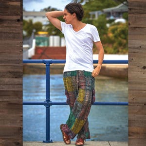 May include: Multi-colored patchwork pants with cargo pockets. The pants feature a mix of green, yellow, and purple hues. The person is wearing a white v-neck t-shirt and brown sandals. The background includes a blue railing and water.