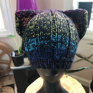 May include: A black knitted hat with cat ears, displaying a colourful rainbow gradient. The hat transitions from purple at the brim to blue, green, yellow, and pink at the ears. The hat is displayed on a black mannequin head.