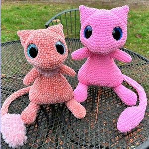 May include: Two handmade crochet plush toys in the shape of a cat-like creature. One is light pink, the other is a brighter pink. Both have large, round, blue eyes and small arms, legs, and tails. The plush toys are sitting on a black metal table.