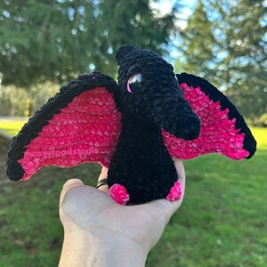 May include: A handmade black and pink crochet pterodactyl with a textured, plush appearance. The wings are a vibrant pink with black edging. The pterodactyl has small pink feet and a detailed head. The text "mossyloodstools" is visible on the wing.