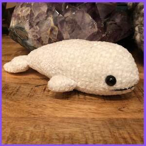 May include: White plush toy shaped like a beluga whale with black eyes. The toy is lying on a wooden surface.