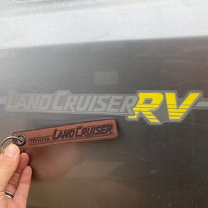 May include: A brown leather Land Cruiser keyring with a silver ring. The keyring features the Toyota and Land Cruiser logos. A Land Cruiser RV sticker is also visible, with yellow and grey lettering.