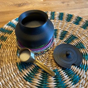 May include: A black ceramic jar with a lid, a small gold spoon filled with white sugar, and a woven straw mat.