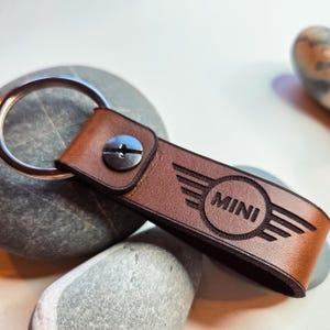 May include: A brown leather MINI keyring with a silver key ring. The keyring features the word "MINI" within a circle, flanked by wing designs. A silver screw fastens the leather strap to the ring.