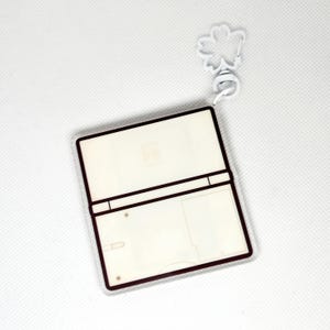 Op de afbeelding: Witte plastic sleutelhanger in de vorm van een gameconsole met een bruine rand. De sleutelhanger heeft een lege ruimte voor een foto of ontwerp.