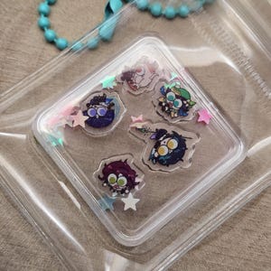 Puede incluir: Una funda cuadrada transparente que contiene seis pegatinas de personajes coloridos y confeti en forma de estrella. Las pegatinas presentan personajes de estilo caricatura con varios colores de pelo y expresiones. La funda está en una bolsa de plástico transparente.