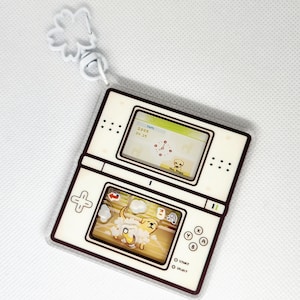 Op de afbeelding: Een witte en bruine plastic sleutelhanger in de vorm van een Nintendo DS handheld-spelconsole. Het bovenste scherm toont een spel met een hondenpersonage. Het onderste scherm toont een hond die wordt gewassen. De sleutelhanger heeft een bloemvormige hanger.