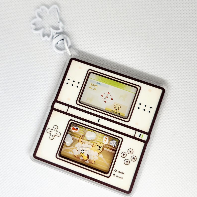 Op de afbeelding: Een witte en bruine plastic sleutelhanger in de vorm van een Nintendo DS handheld-spelconsole. Het bovenste scherm toont een spel met een hondenpersonage. Het onderste scherm toont een hond die wordt gewassen. De sleutelhanger heeft een bloemvormige hanger.