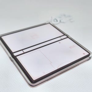 Op de afbeelding: Een witte en bruine plastic sleutelhanger in de vorm van een draagbare gameconsole. De sleutelhanger heeft een leeg scherm en knoppen.