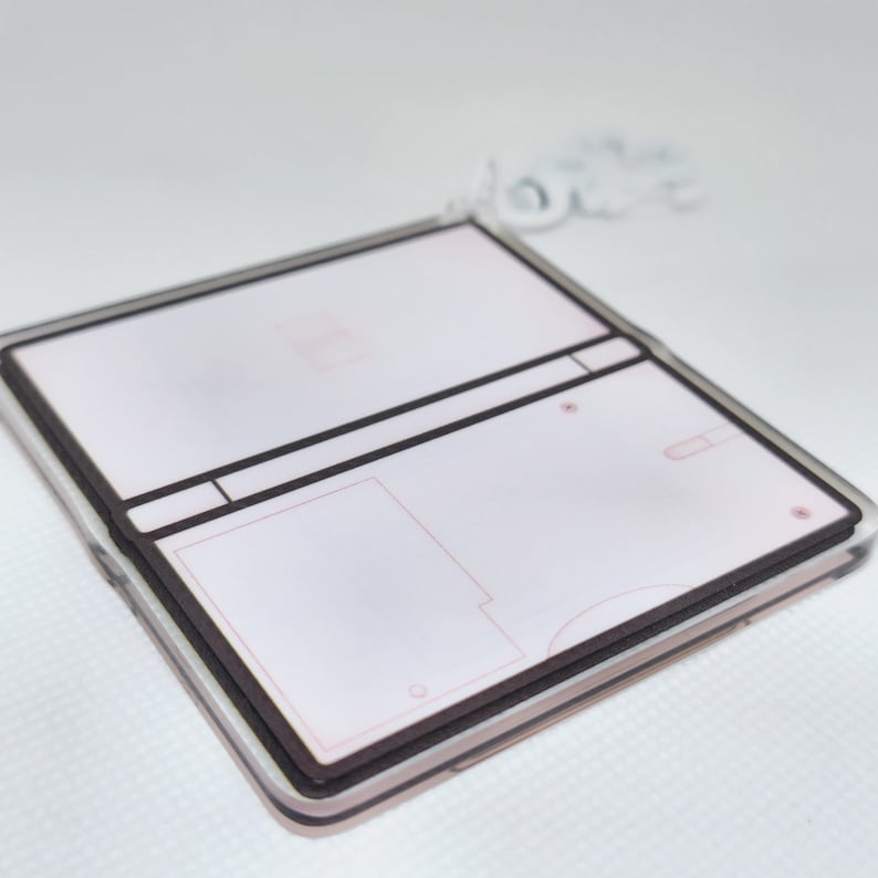 Op de afbeelding: Een witte en bruine plastic sleutelhanger in de vorm van een draagbare gameconsole. De sleutelhanger heeft een leeg scherm en knoppen.