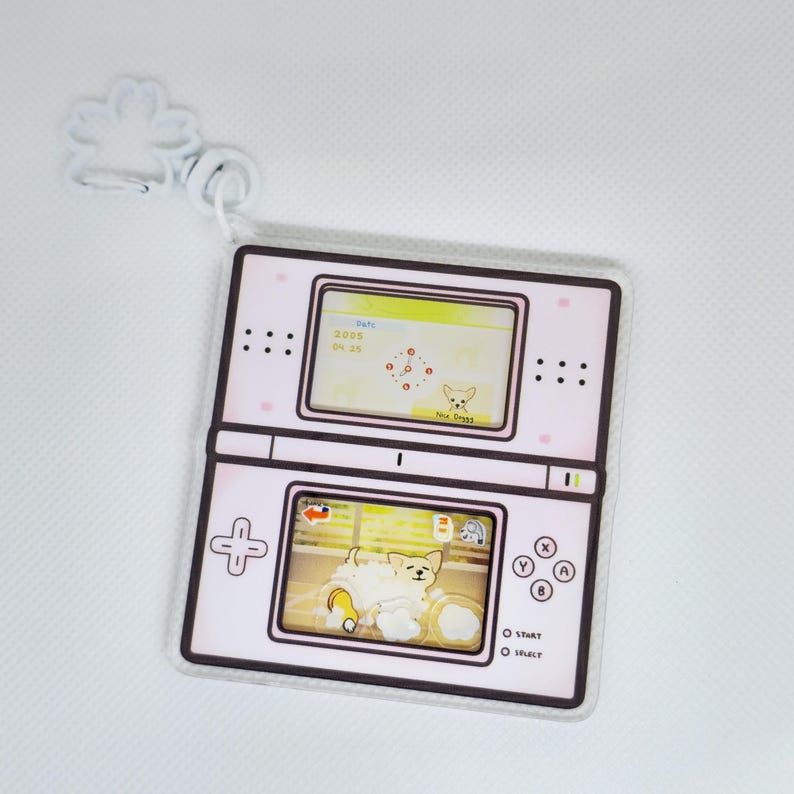 Op de afbeelding: Een roze en witte sleutelhanger in de vorm van een Nintendo DS handheld-spelconsole. Het scherm toont een spel met een cartoonhond die een bad neemt. De sleutelhanger heeft een kleine metalen lus om aan een tas of sleutels te bevestigen.