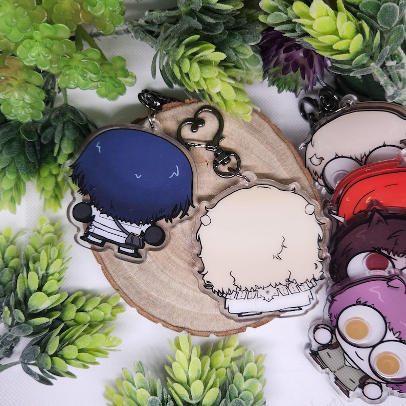 Op de afbeelding: Transparante acryl sleutelhangers met anime-stijl personages met verschillende kapsels en outfits. De sleutelhangers zijn bevestigd aan een zilverkleurige sluiting. De achtergrond bevat groen en een houten schijf.