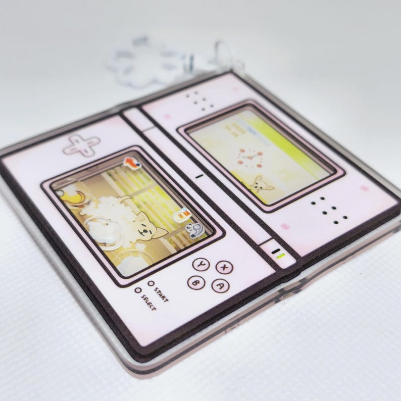 Op de afbeelding: Een roze en witte handheld-videogamesysteem met een scherm waarop een cartoonhond te zien is. De console heeft knoppen met de labels "Start", "Select", "A", "B", "X", "Y" en een directionele pad.