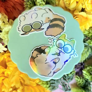 May include: Two holographic stickers featuring cartoon cats with large circular eyes. The top sticker shows a white and brown cat with a striped tail. The bottom sticker depicts a calico cat with patches of brown, black, and white fur.