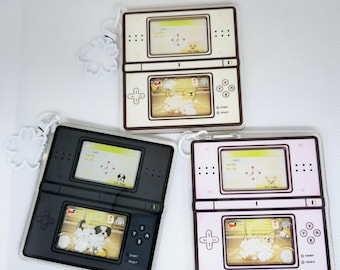 May include: Three keychain charms shaped like a Nintendo DS handheld gaming console. Each charm features a different color scheme: white, black, and pink. The charms have a screen displaying a dog game with the text "Pet Puppy".