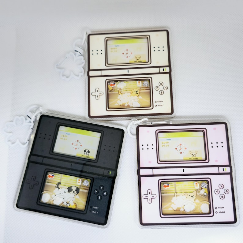 Op de afbeelding: Drie sleutelhangers in de vorm van een Nintendo DS handheld-spelconsole. Elke hanger heeft een ander kleurenschema: wit, zwart en roze. De hangers hebben een scherm dat een hondenspeelgoed met de tekst "Pet Puppy" weergeeft.